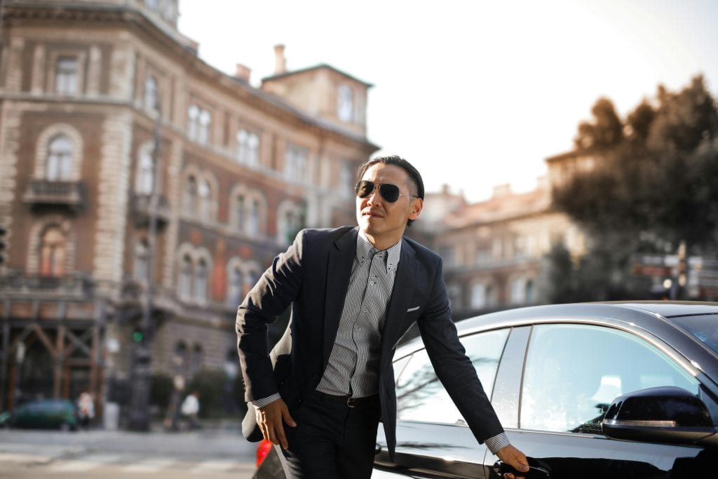 Confident businessman in sunglasses standing by car in city street.