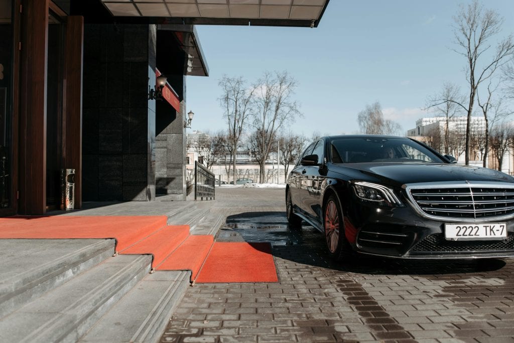 A luxury black sedan parked on a red carpet driveway outside a modern building.