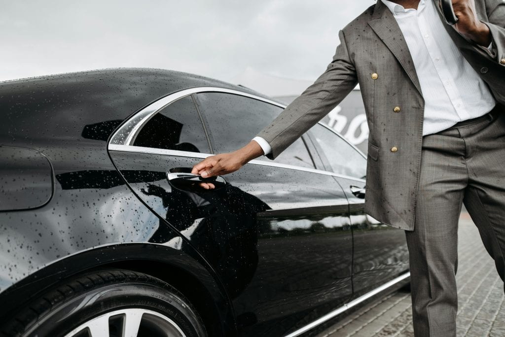 A man in a suit opens a luxury car door under raindrops on stone pavement.