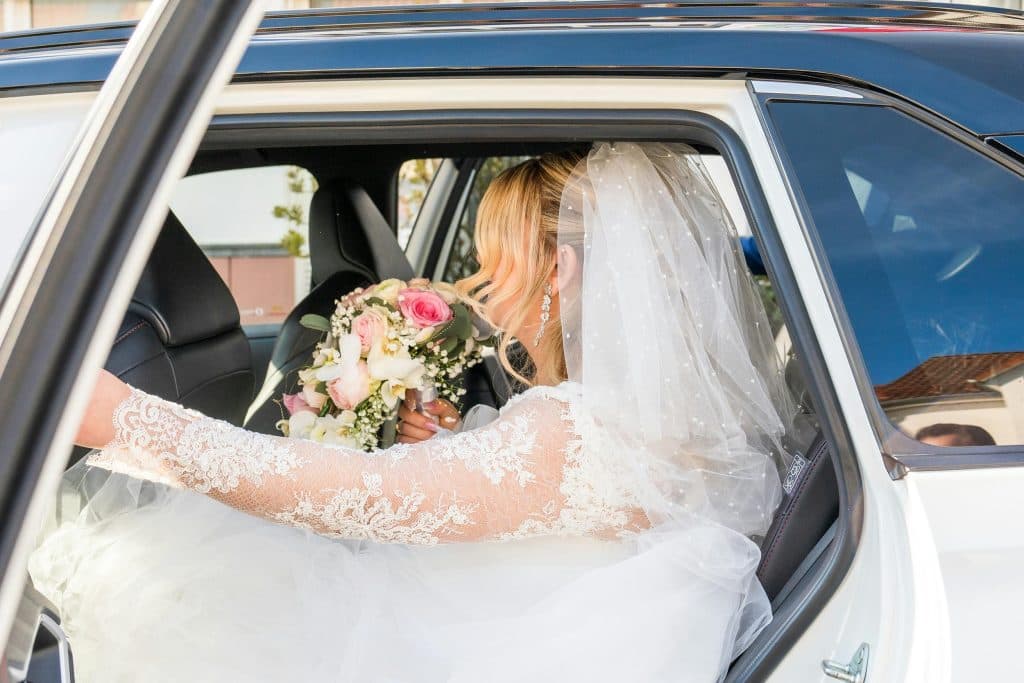 Elegant bride in lace wedding dress and veil holding a bouquet inside a car.