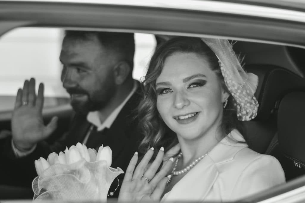 Elegant black and white portrait of a bride and groom waving from a car, celebrating their wedding day.