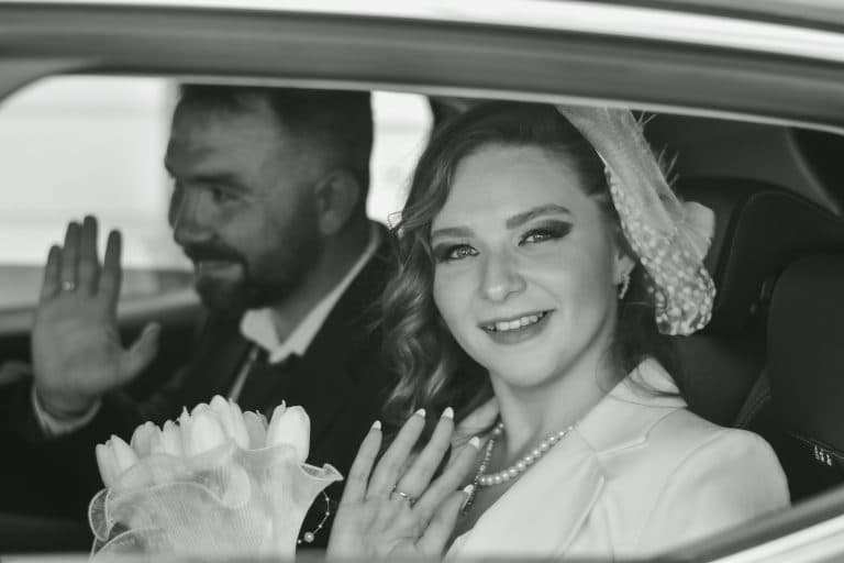 Elegant black and white portrait of a bride and groom waving from a car, celebrating their wedding day.