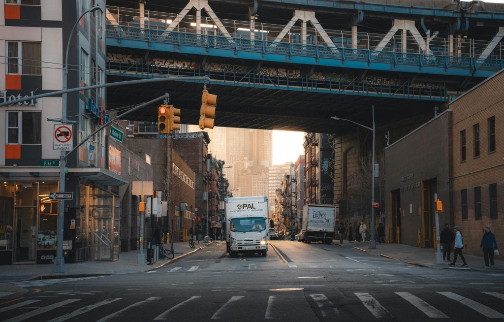 A typical street scene in New York City with trucks under a bridge and pedestrians walking.