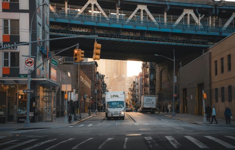 A typical street scene in New York City with trucks under a bridge and pedestrians walking.