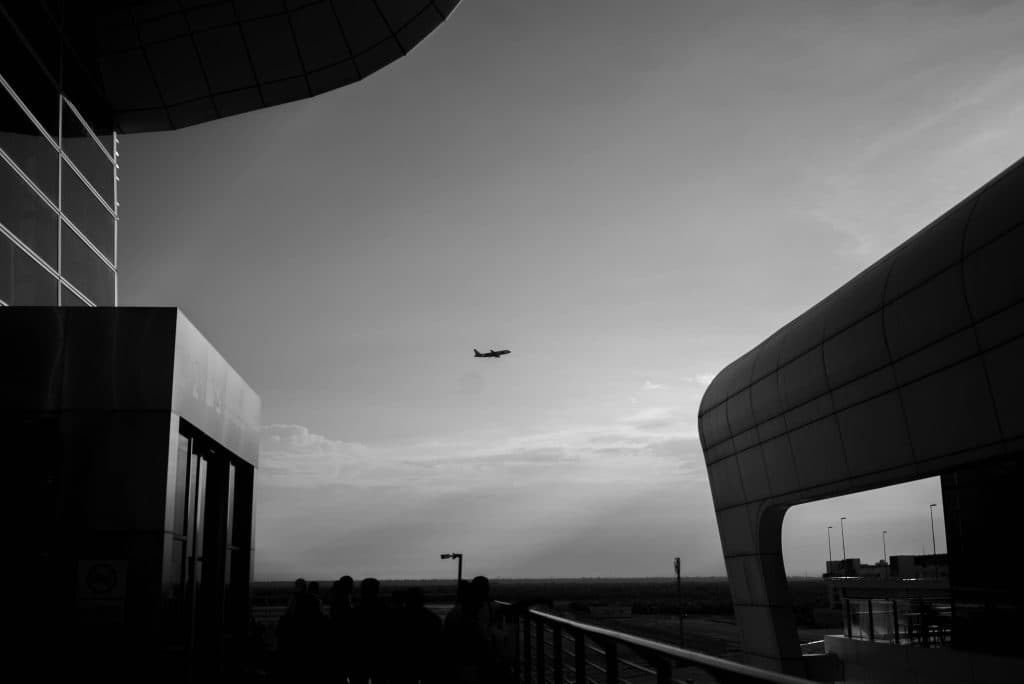 Black and white photo of an airplane flying above a modern airport terminal at sunset.