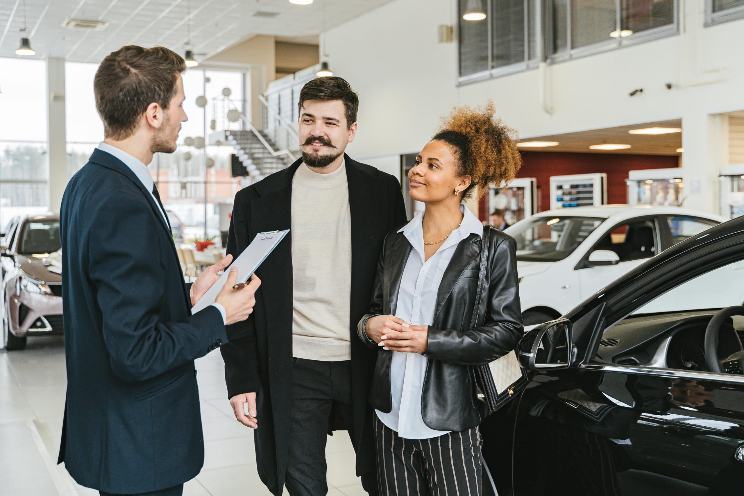 Blogs | Articles | News | Tips & Tricks | Video | FAQ | Infomation 12 Couple discussing car purchase with salesman at indoor dealership, engaging conversation.