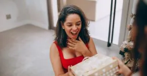 A woman in a red dress excitedly opens a beautifully wrapped Christmas gift indoors.