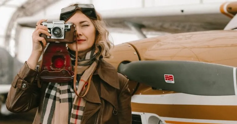 Fashionable woman with vintage camera posing by an airplane in an open hangar.