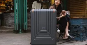 Asian woman at café holding coffee, seated with suitcase in urban setting.