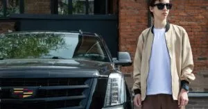 Young man in sunglasses and jacket standing next to a Cadillac Escalade against a brick building.