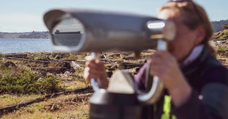 A woman uses a coin-operated binocular to view a distant ship along the Colwood shores.