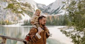 Father carrying his child by a serene lake amidst majestic mountains, enjoying nature.