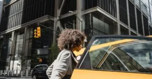 Confident woman with afro hairstyle getting into a city taxi by modern architecture.