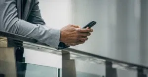 Crop anonymous black businessman in formal suit leaning on fence and using phone in soft daylight