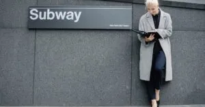 Full body smiling young businesswoman in formal clothes and coat reading documents and standing near subway station entrance