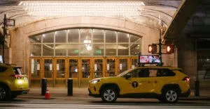 A yellow taxi cab parked outside a well-lit building entrance at night in an urban setting.