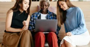 Three diverse young women collaborating on a laptop, indoors, in a modern workspace.