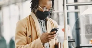 Stylish man with dreadlocks wearing a face mask on public transportation, checking his phone. Urban lifestyle context.