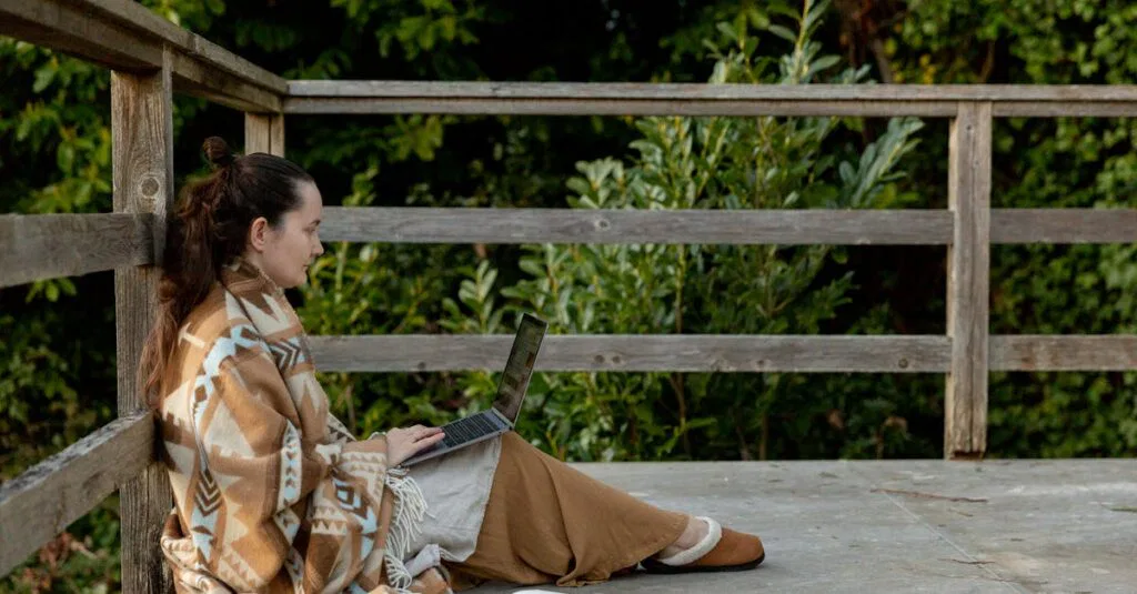 A woman wrapped in a blanket works on her laptop outside on a terrace, enjoying a tranquil autumn afternoon.