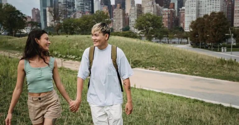 Positive diverse couple in casual apparel holding hands while walking on grass lawn near contemporary buildings and looking at each other
