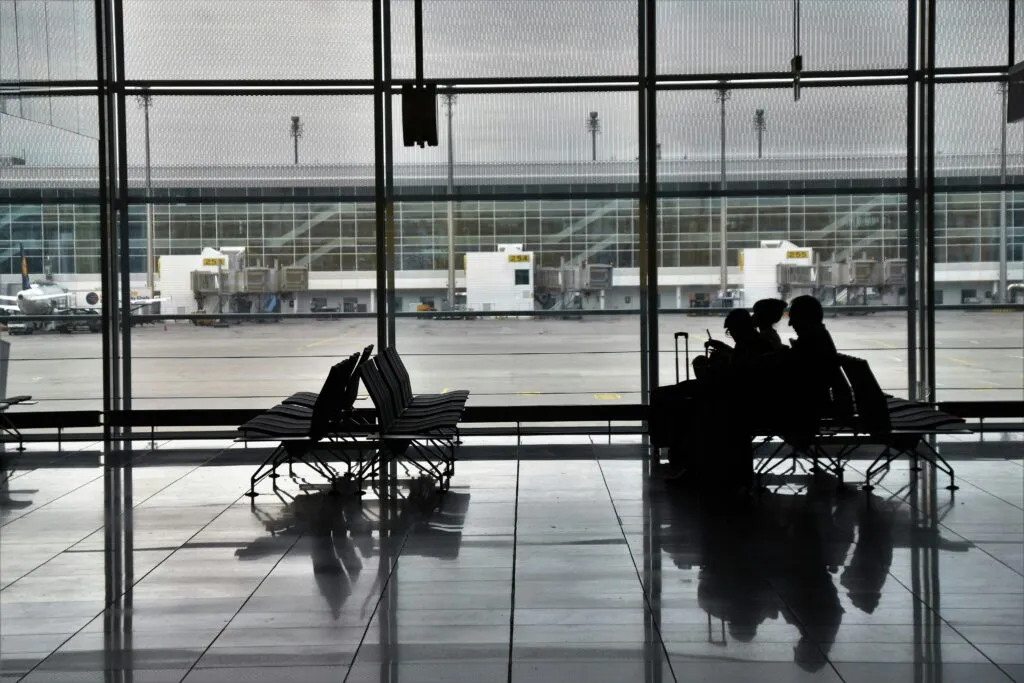 Silhouette of unrecognizable passengers sitting on chairs with luggage near window and waiting for flight