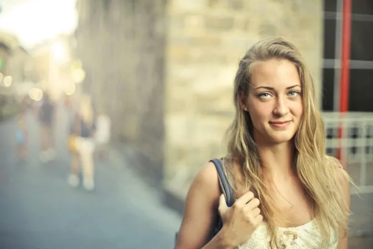 Portrait of a smiling blonde woman carrying a bag, with a bright and cheerful expression in an urban setting.