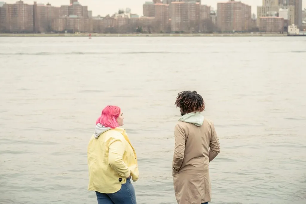 Two friends enjoying a peaceful moment by the riverbank with a cityscape in the background.