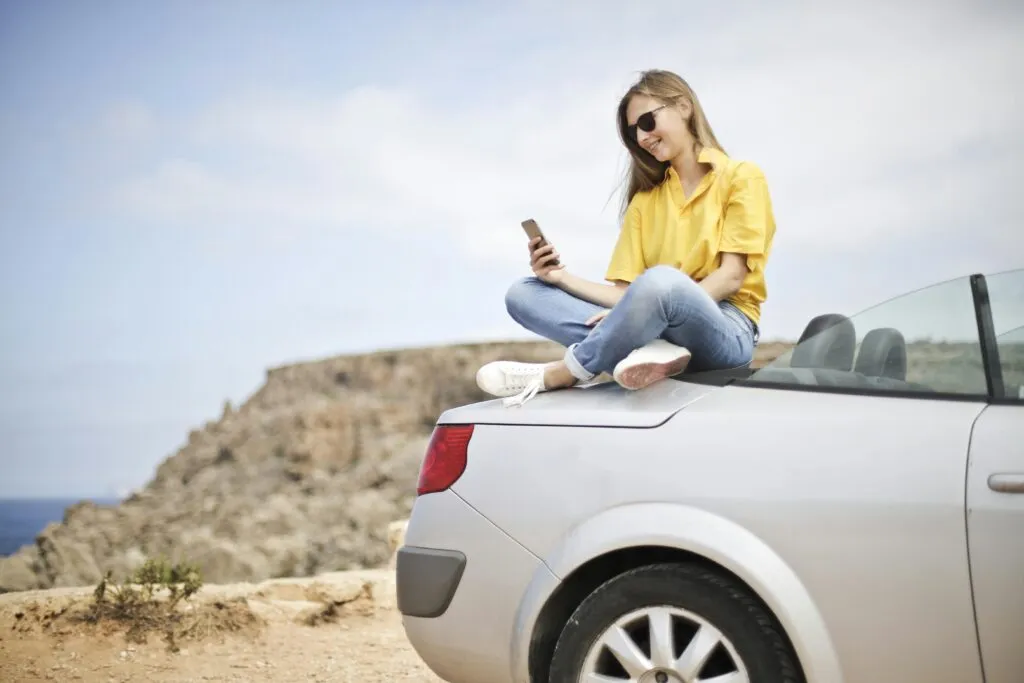 Epic Car Services NYC for Events: The Insider’s Guide to Flawless Rides in 2025 9 Smiling young woman sitting on a car at the beach in Mgarr, Malta using a smartphone.