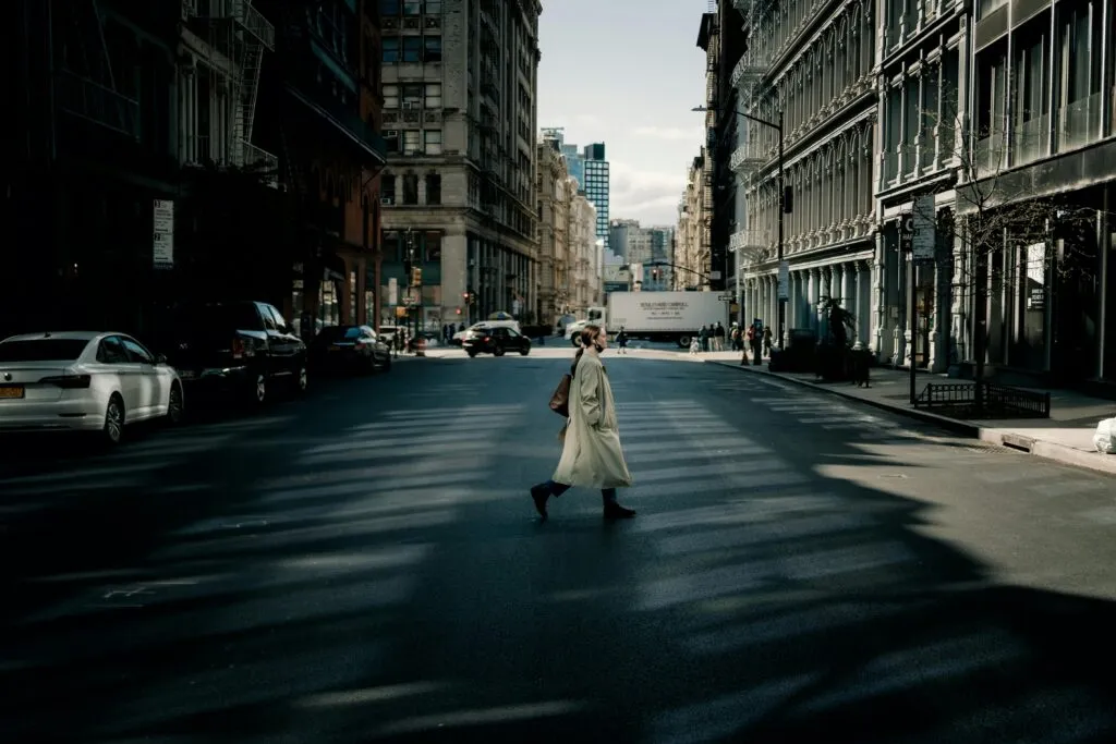 A woman in a trench coat walks across a sunlit street in New York City with tall buildings surrounding her.