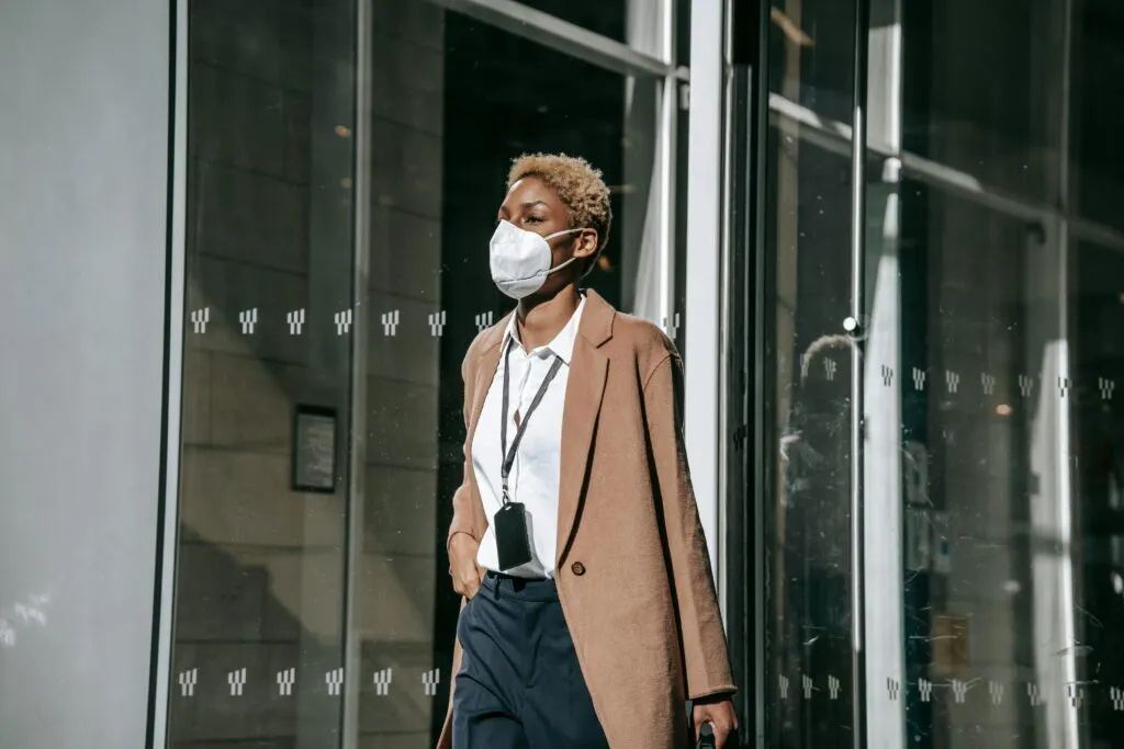 Contemplative young African American businesswoman in formal clothes and face mask walking along modern building glass wall on sunny autumn day