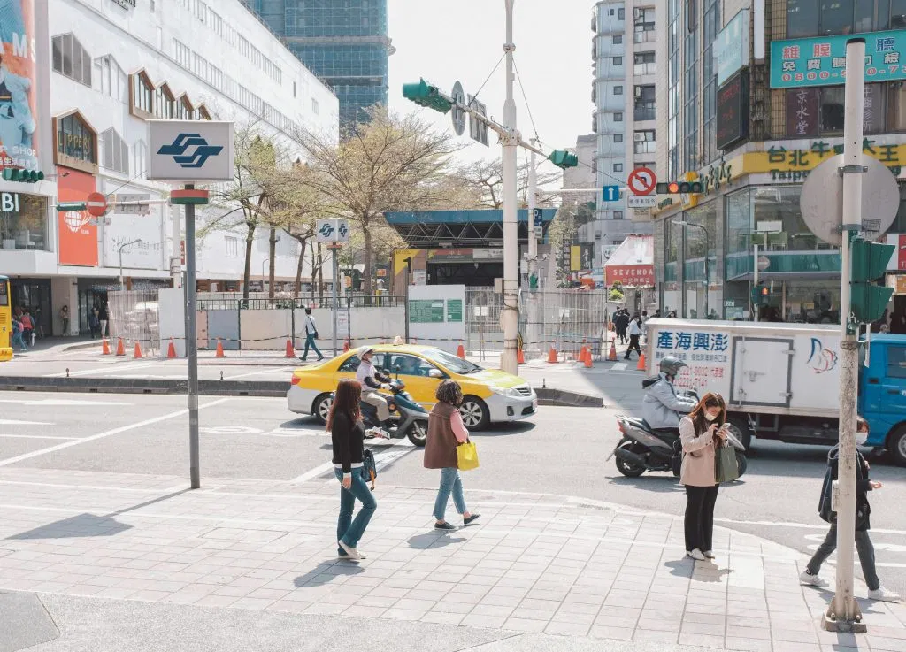 People wearing masks crossing a busy city intersection on a sunny day, with traffic and buildings in the background.