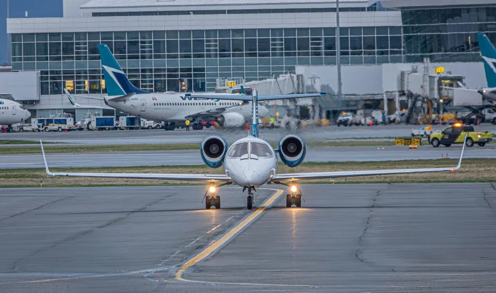 A private jet on the runway at a bustling airport with commercial planes and terminal in view.